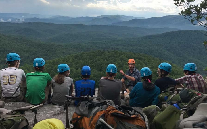 A group of students wearing helmets sit in a line and listen to an instructor speak. In the background, there is a vast and green mountainous landscape.